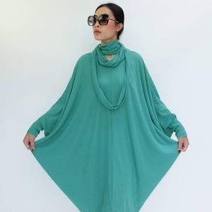 K&ouml;nnte beinhalten: Ein teal-farbenes Langarmkleid mit einem einzigartigen, flie&szlig;enden Design. Das Kleid hat einen Wasserfallausschnitt und einen Schlitz am Saum. Die Person auf dem Bild tr&auml;gt eine gro&szlig;e, quadratische Sonnenbrille.