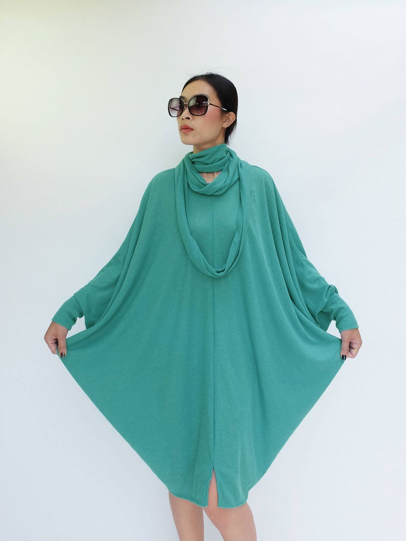 K&ouml;nnte beinhalten: Ein teal-farbenes Langarmkleid mit einem einzigartigen, flie&szlig;enden Design. Das Kleid hat einen Wasserfallausschnitt und einen Schlitz am Saum. Die Person auf dem Bild tr&auml;gt eine gro&szlig;e, quadratische Sonnenbrille.