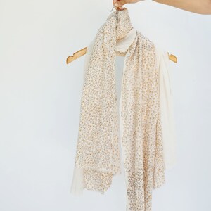 May include: A white scarf with a delicate floral pattern in gold. The scarf is hanging on a wooden hanger.