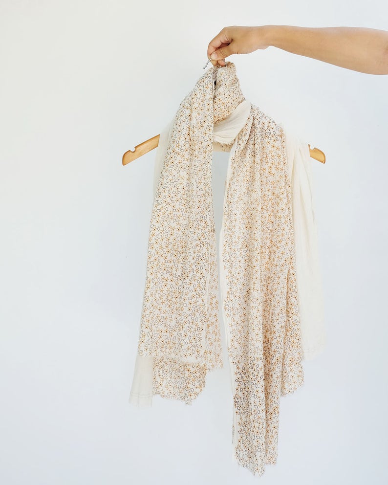 May include: A white scarf with a delicate floral pattern in gold. The scarf is hanging on a wooden hanger.
