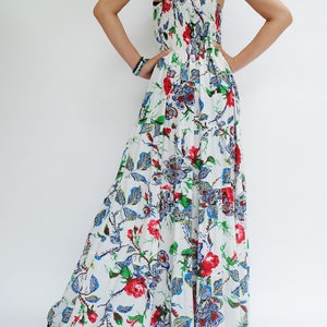 May include: White maxi dress with a floral pattern featuring red roses, blue and green leaves. The dress has thin straps and a tiered skirt. The back of the dress is visible, with a gathered bodice and a cutout detail.