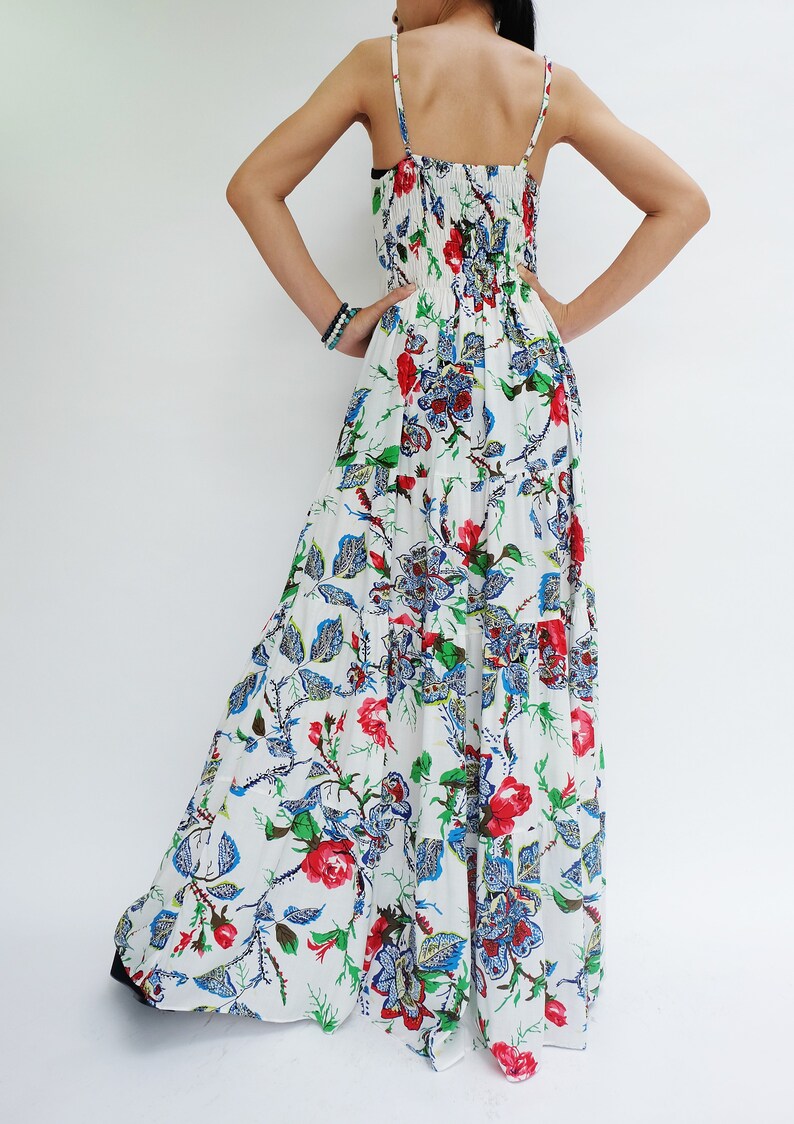 May include: White maxi dress with a floral pattern featuring red roses, blue and green leaves. The dress has thin straps and a tiered skirt. The back of the dress is visible, with a gathered bodice and a cutout detail.