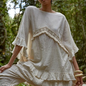May include: A woman wearing a white, loose-fitting top with a crocheted trim and white pants. She is standing in a forest setting.