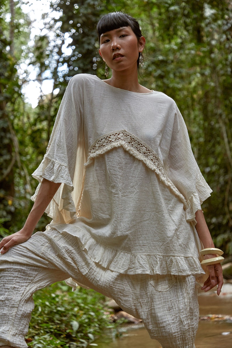 May include: A woman wearing a white, loose-fitting top with a crocheted trim and white pants. She is standing in a forest setting.