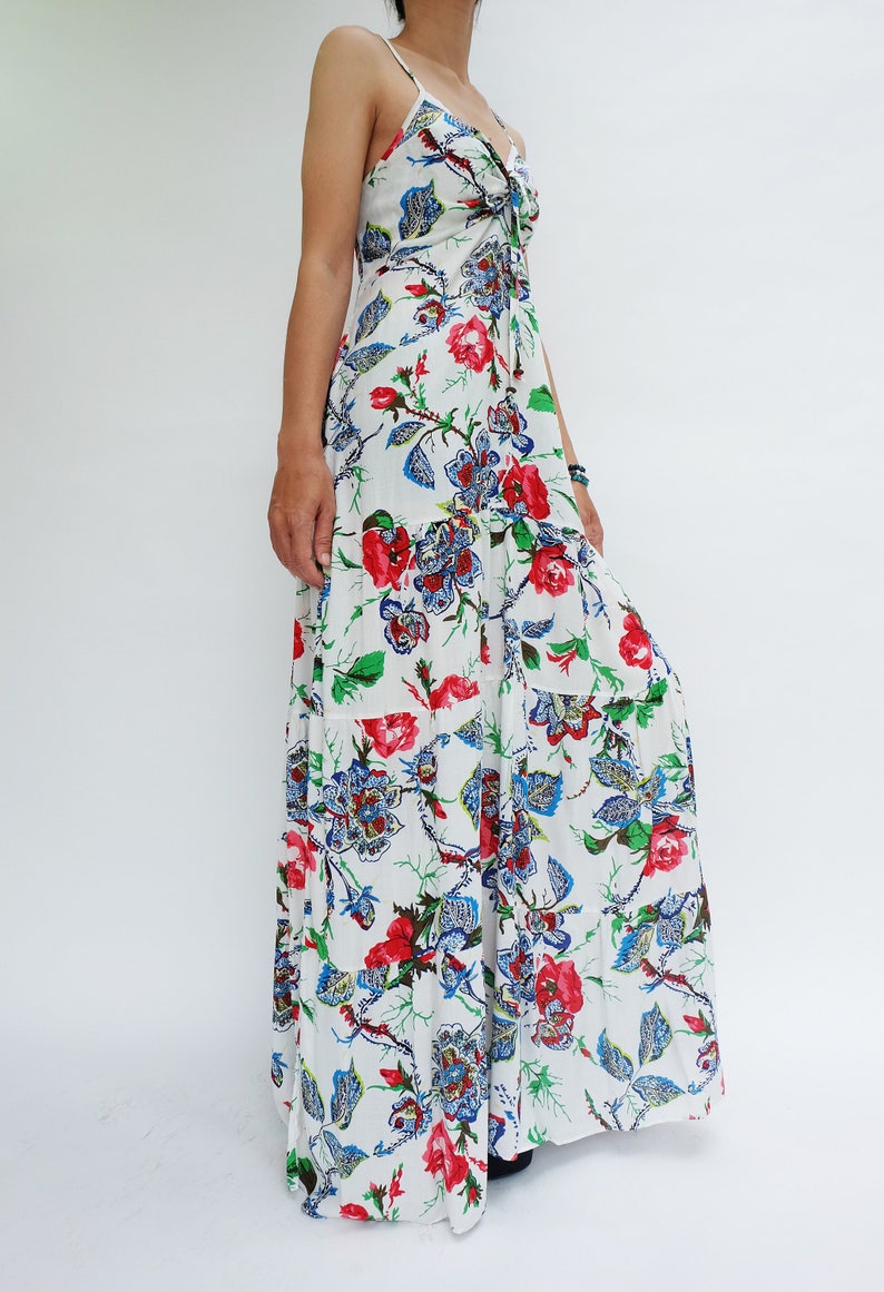 May include: A white maxi dress with a floral pattern featuring red, blue, and green accents. The dress has spaghetti straps and a tiered skirt. The floral design includes roses and other stylized flowers.