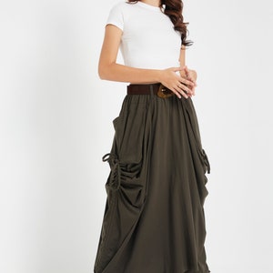 Puede incluir: Una mujer lleva una camiseta blanca ajustada y una falda larga de color verde oscuro con un cintur&oacute;n marr&oacute;n y una hebilla grande. La falda tiene un corte holgado y abullonado y presenta un detalle de cord&oacute;n en los laterales.