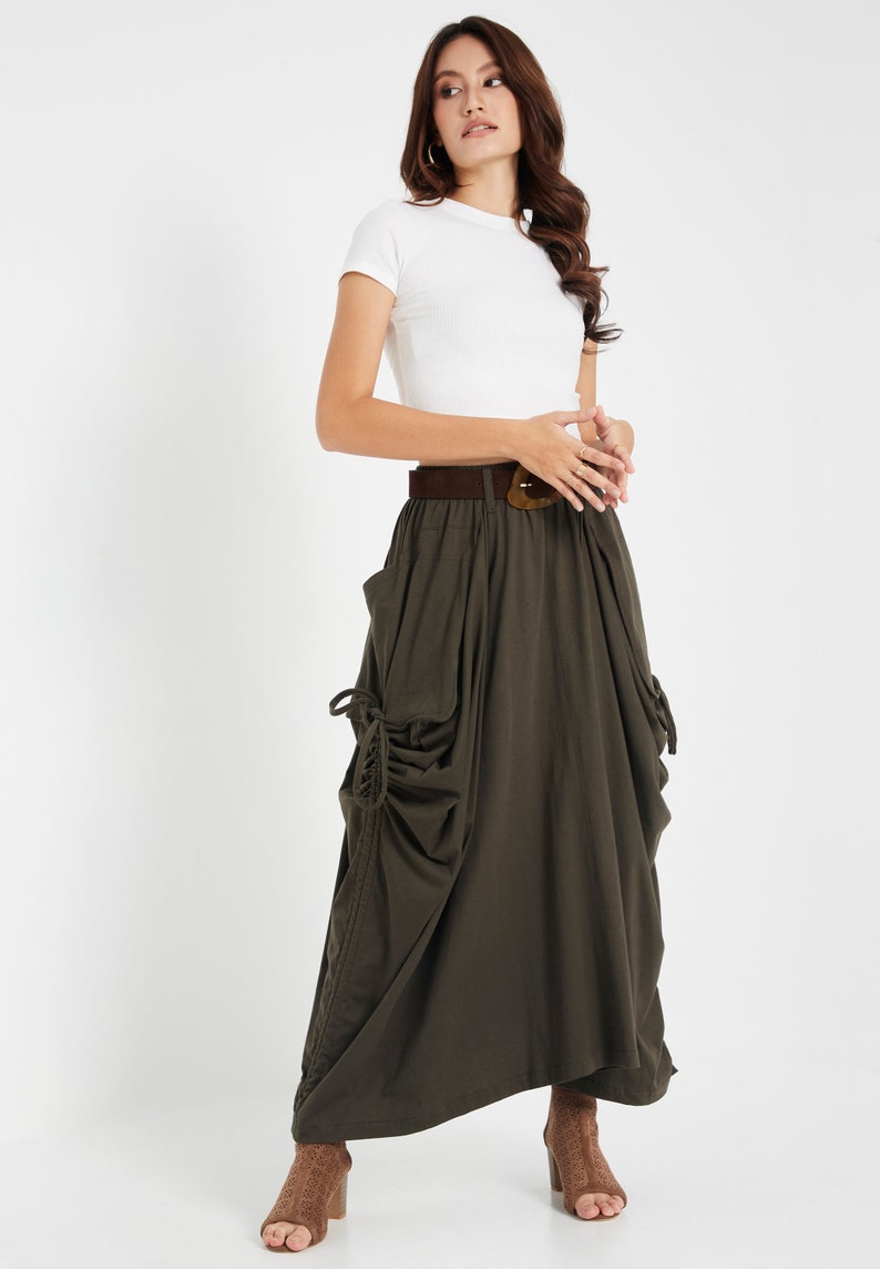 Puede incluir: Una mujer lleva una camiseta blanca ajustada y una falda larga de color verde oscuro con un cintur&oacute;n marr&oacute;n y una hebilla grande. La falda tiene un corte holgado y abullonado y presenta un detalle de cord&oacute;n en los laterales.