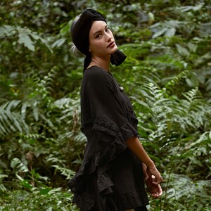 K&ouml;nnte beinhalten: Eine Frau tr&auml;gt ein schwarzes, gerafftes Oberteil mit Spitzendetails und einen schwarzen Turban. Sie steht in einer &uuml;ppigen gr&uuml;nen Waldlandschaft.