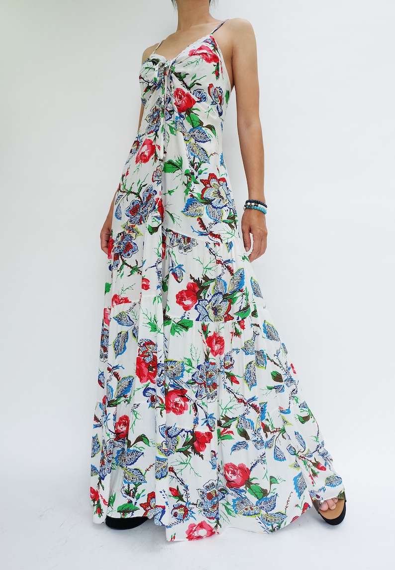 May include: White maxi dress with a floral print featuring red roses, blue and green leaves. The dress has spaghetti straps and a tie detail at the bust. The tiered skirt flows to the floor, creating a bohemian style.