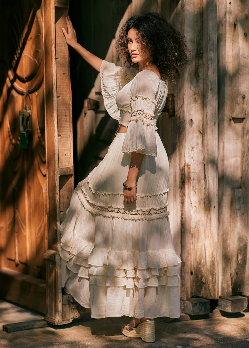 Puede incluir: Una mujer con una falda maxi blanca de capas con ribete de encaje y un top blanco a juego con detalles de encaje. Est&aacute; de pie frente a una puerta de madera.