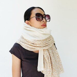 May include: A woman wearing a white and floral patterned scarf with a white top and black t-shirt. She is wearing black sunglasses.