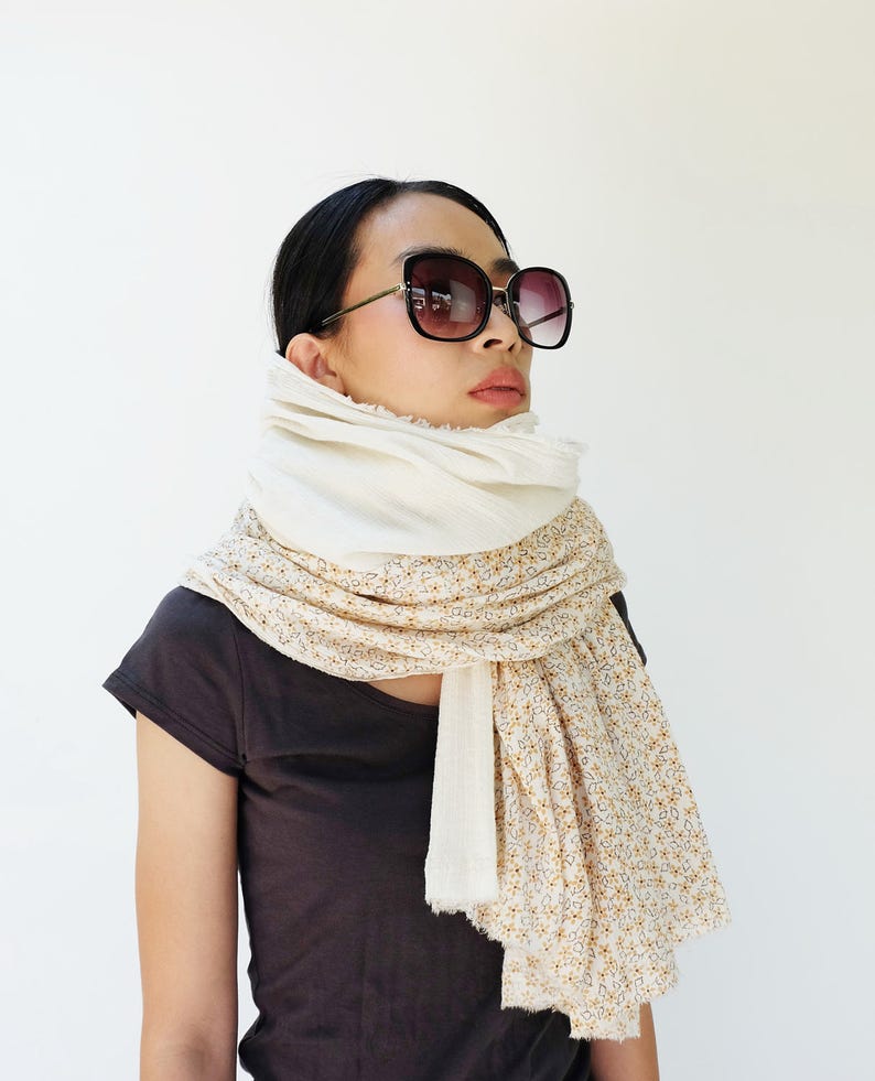 May include: A woman wearing a white and floral patterned scarf with a white top and black t-shirt. She is wearing black sunglasses.