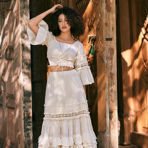 Puede incluir: Una mujer con un top blanco estilo campesino, de hombros descubiertos, con una falda con volantes. El top tiene un lazo en la cintura y la falda tiene varias capas de volantes. Est&aacute; de pie frente a una puerta de madera.