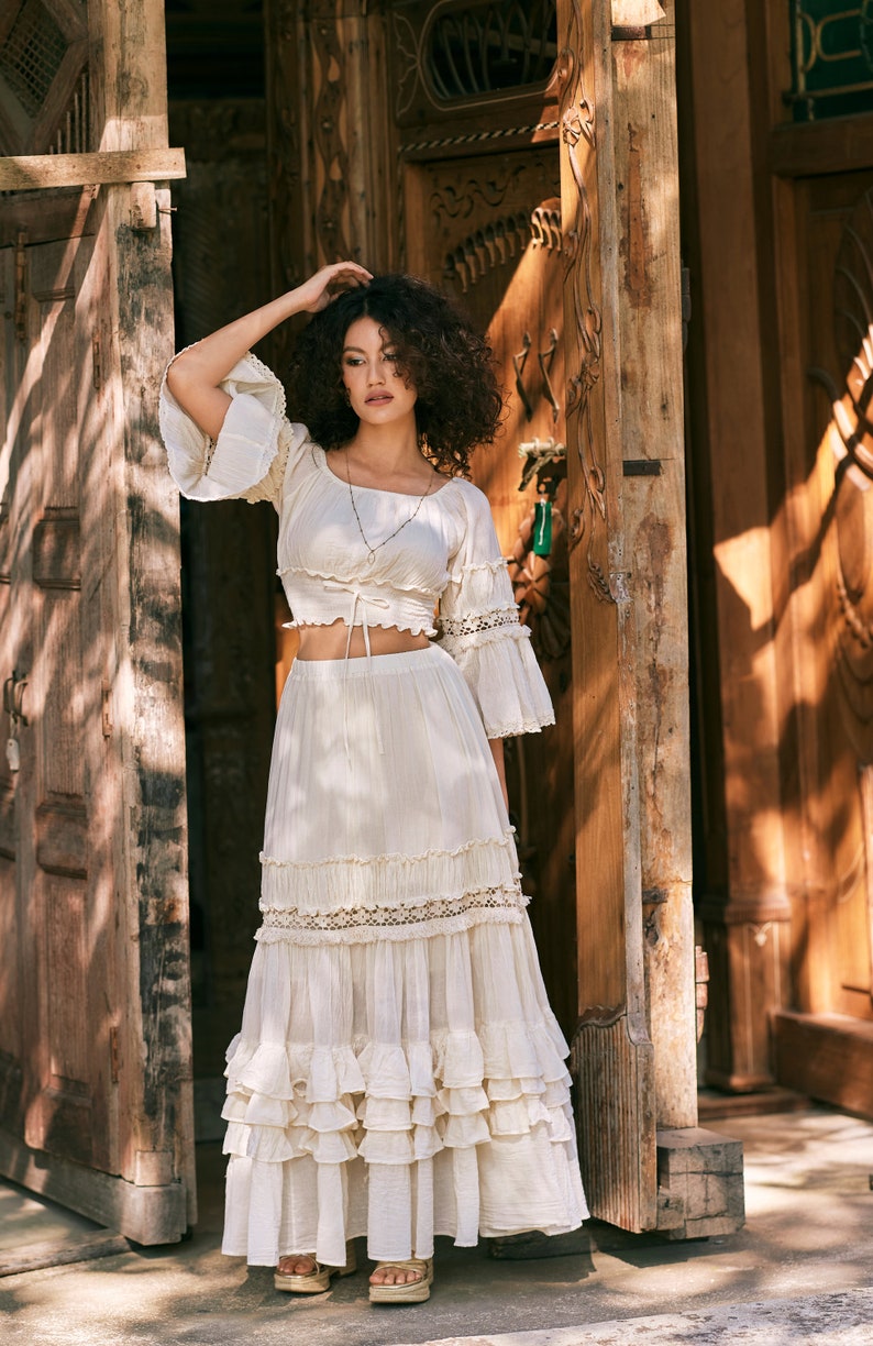 Puede incluir: Una mujer con un top blanco estilo campesino, de hombros descubiertos, con una falda con volantes. El top tiene un lazo en la cintura y la falda tiene varias capas de volantes. Est&aacute; de pie frente a una puerta de madera.