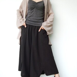 May include: A woman wearing a long, flowy, light brown cardigan over a gray tank top and a black, wide-legged, asymmetrical skirt with pockets. She is wearing black sandals with cork wedges.