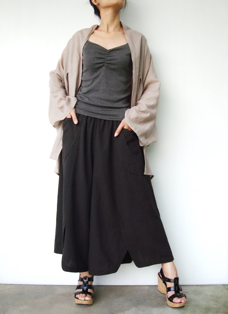May include: A woman wearing a long, flowy, light brown cardigan over a gray tank top and a black, wide-legged, asymmetrical skirt with pockets. She is wearing black sandals with cork wedges.