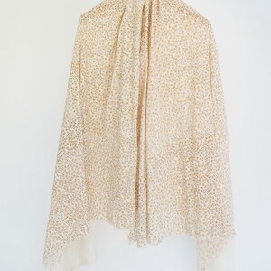 May include: A white scarf with a small, gold floral pattern. The scarf is hanging on a hanger.
