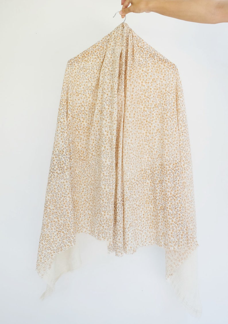 May include: A white scarf with a small, gold floral pattern. The scarf is hanging on a hanger.