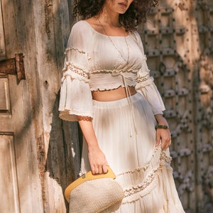 Puede incluir: Una mujer con un top y una falda de crochet blanco con un bolso de mano tejido amarillo. El top tiene un lazo en la cintura y la falda tiene un dobladillo con volantes.