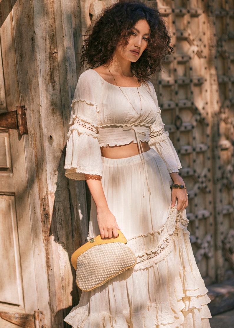 Puede incluir: Una mujer con un top y una falda de crochet blanco con un bolso de mano tejido amarillo. El top tiene un lazo en la cintura y la falda tiene un dobladillo con volantes.
