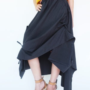 May include: A black asymmetrical skirt with a high waist and a tie detail at the side. The skirt is made of a soft, flowing fabric and is paired with brown wedge sandals.