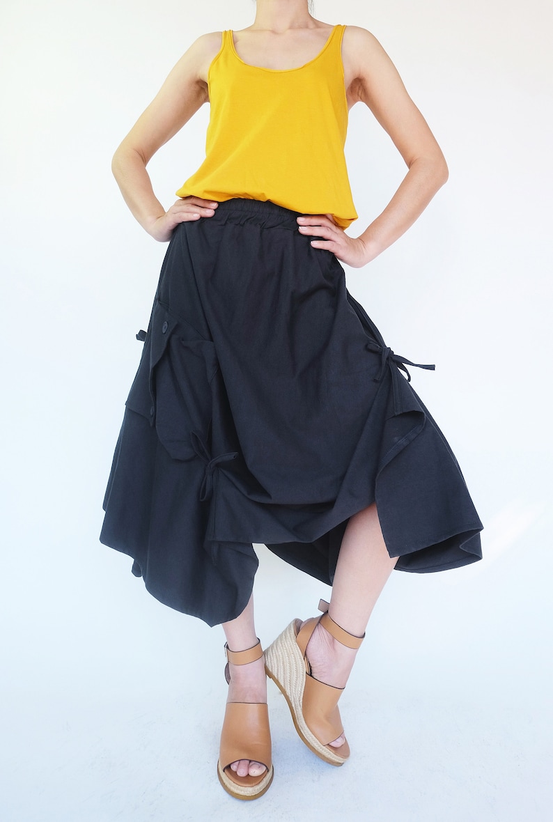 May include: A woman is wearing a yellow tank top and a black asymmetrical skirt with pockets and ties. She is wearing brown wedge sandals.
