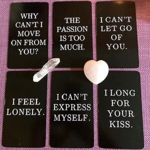 Op de afbeelding: Zes rechthoekige zwarte kaarten met witte tekst: WHY CAN'T I MOVE ON FROM YOU?, THE PASSION IS TOO MUCH., I CAN'T LET GO OF YOU., I FEEL LONELY., I CAN'T EXPRESS MYSELF. en I LONG FOR YOUR KISS. Een heldere kwartspunt en een lichtroze hartvormige steen.