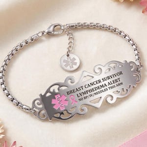 May include: A silver-toned medical alert bracelet featuring a box chain and a decorative filigree plate. The plate is engraved with BREAST CANCER SURVIVOR, LYMPHEDEMA ALERT, and NO BP/IV/NEEDLES THIS ARM, alongside a pink medical symbol and ribbon.
