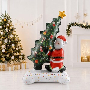 May include: A large inflatable decoration featuring a green Christmas tree with ornaments and a gold star on top, held by a smiling Santa Claus figure in a red suit. The entire structure stands on a white base decorated with colorful stars.
