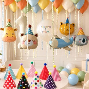 May include: A collection of party decorations featuring animal-shaped balloons including a giraffe, bear, tiger, and koala, alongside a blue airplane balloon and a clear balloon with the text "САВЕЛИЮ 1 ГОДИК". Colorful party hats are arranged on a table.