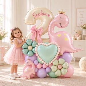 May include: A large balloon arrangement featuring a cream-colored number two balloon with the name Sophia printed in pink, a pink dinosaur balloon with a gold crown, a mint green heart balloon, two flower-shaped balloons, and various pastel balloons.