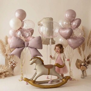 Puede incluir: Una colección de globos en tonos rosa empolvado, crema y transparentes con confeti, que incluye un globo grande con forma de lazo y un globo con forma de caballo balancín. Los globos están dispuestos en racimos con cintas decorativas.