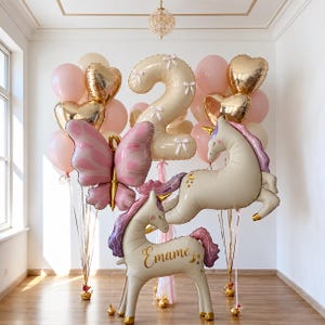 May include: A balloon arrangement featuring a large cream-colored number two balloon with white bows, a pink butterfly balloon, two cream-colored unicorn balloons with purple manes, and gold heart balloons, with the name Emame printed on one.