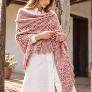 Pode incluir: Um xale de tricô em tom rosa seco com um padrão texturizado de ponto waffle e franjas longas nas bordas. O xale está drapeado sobre uma camisa branca de botões, destacando o trabalho complexo do fio e a cor rosa suave e fosca.