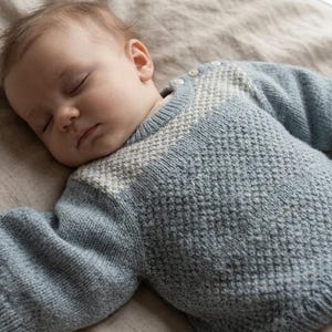 Könnte beinhalten: Ein langärmeliger Strickpullover in einem sanften Blauton mit einem strukturierten Rautenmuster an Brust und Schultern. Das Kleidungsstück hat einen Rundhalsausschnitt mit zwei kleinen weißen Knöpfen an der Schulternaht sowie gerippte Bündchen.