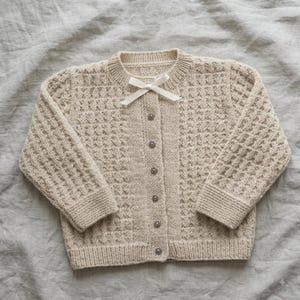 Könnte beinhalten: Eine beigefarbene Strickjacke mit strukturiertem Waffelmuster, langen Ärmeln und einem gerippten Rundhalsausschnitt. Der Vorderverschluss besteht aus sechs kleinen runden Knöpfen und einer dekorativen weißen Schleife am Kragen.
