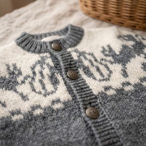 Könnte beinhalten: Eine grau-weiße Strickjacke mit Rundhalsausschnitt, einer Knopfleiste mit drei metallischen Knöpfen und einem sich wiederholenden Muster aus stilisierten Tierfiguren auf Brust und Schultern, gefertigt mit geripptem Kragen und Knopfleiste.