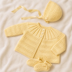 Könnte beinhalten: Ein hellgelbes, gestricktes Baby-Set bestehend aus einer langärmeligen Strickjacke mit plissierter Passe und drei Knöpfen, einer passenden Haube mit Satinband sowie einem Paar kleiner Strickschühchen mit dekorativen Schleifen.