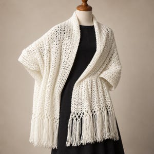 May include: An off-white knitted shawl featuring an openwork stitch pattern and long fringe along the bottom edge. The shawl is draped over a black garment on a mannequin, showcasing the textured yarn construction and the vertical drape.