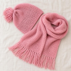 Könnte beinhalten: Eine weiche, rosa Strickmütze mit einem großen, passenden Bommel oben, kombiniert mit einem langen, rechteckigen Strickschal, der an beiden Enden Fransen aufweist. Beide Artikel sind aus einem leichten Garn mit offener Webstruktur gefertigt.
