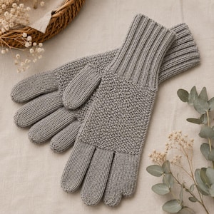 May include: A pair of grey knit gloves featuring a textured seed stitch pattern on the back of the hand and fingers, complemented by a ribbed cuff design that extends up the wrist for a secure fit. The gloves are crafted with a consistent grey yarn.