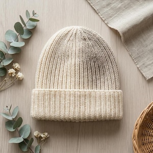 Könnte beinhalten: Eine cremefarbene Strickmütze mit gerippter Textur und einem breiten, umgeschlagenen Saum. Die Mütze weist ein vertikales Strickmuster über die gesamte Krone und den Rand auf und zeigt eine gleichmäßige, cremeweiße Garnstruktur mit weicher Optik.