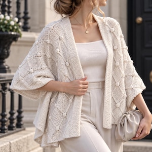 May include: An off-white open-front cardigan featuring a textured diamond knit pattern with small raised bobbles at each intersection. The garment has wide, loose sleeves and a relaxed drape, worn over a matching ribbed tank top and trousers.