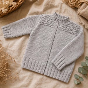 Könnte beinhalten: Eine hellgraue, langärmelige Strickjacke mit einem strukturierten Rautenmuster im oberen Brust- und Schulterbereich, einem gerippten Stehkragen sowie gerippten Bündchen an Ärmeln und Saum. Das Kleidungsstück hat eine vertikale Mittelnaht.
