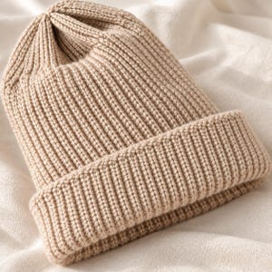 Peut inclure: Un bonnet en tricot beige présentant une texture côtelée verticale épaisse sur tout le corps et un large revers plié à la base. Le bonnet est fabriqué avec un tissage en laine épaisse et uniforme qui crée une forme de couronne arrondie.