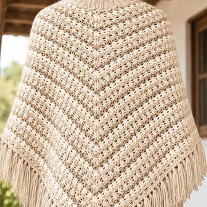 Pode incluir: Um poncho de crochê bege e creme com gola alta canelada e bainha em forma de V. A peça é confeccionada com um padrão de pontos texturizados repetitivos e finalizada com franjas longas e grossas ao longo de toda a borda inferior.