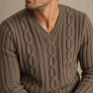 Könnte beinhalten: Ein taupefarbener V-Ausschnitt-Pullover mit einem vertikalen Zopfstrickmuster, das sich mit gerippten Strickspalten abwechselt. Das Kleidungsstück besteht aus einem dicken, strukturierten Strickstoff, der vom Halsausschnitt bis zum Rumpf und den Ärmeln reicht.