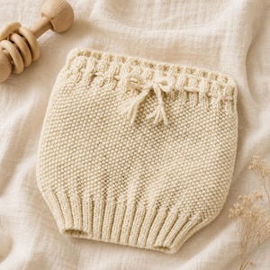 May include: A cream-colored knit diaper cover featuring a textured seed stitch pattern, a ribbed waistband with a decorative woven drawstring tie, and a ribbed leg opening. The garment is crafted with a consistent knit construction throughout.