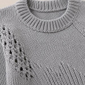 Könnte beinhalten: Ein hellgrauer Strickpullover mit geripptem Rundhalsausschnitt und einem strukturierten Zopfstrickmuster entlang der Schulter und des Ärmels. Das Kleidungsstück ist aus dickem Garn gefertigt und zeigt ein gleichmäßiges Maschenbild.