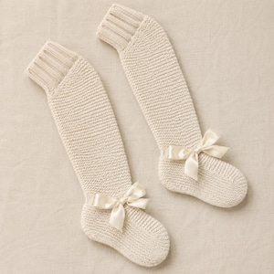 May include: A pair of cream-colored knee-high socks crafted from a textured knit fabric. Each sock features a ribbed cuff at the top and a smooth, matching satin ribbon bow securely attached to the side of the foot area near the toe section.